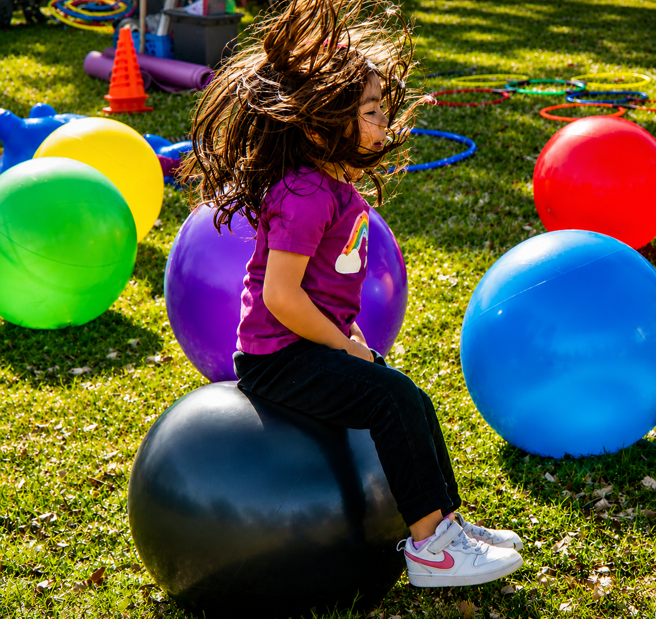 Girl bouncing on a hopper ball with hair flying, colorful balls and hoops in the background at a Sunshine Hoops event